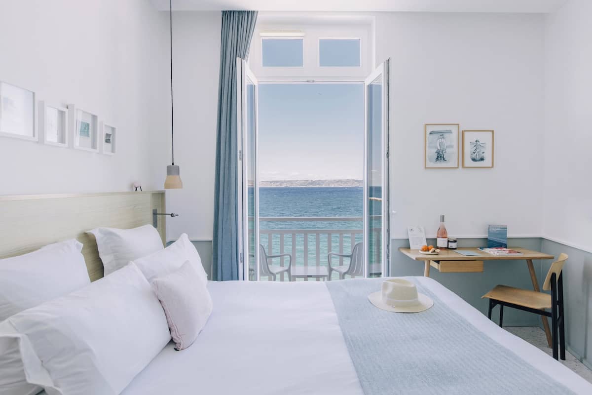 A hotel room with a bed, a desk, a chair, a balcony with a view of the sea, and framed pictures on the wall.