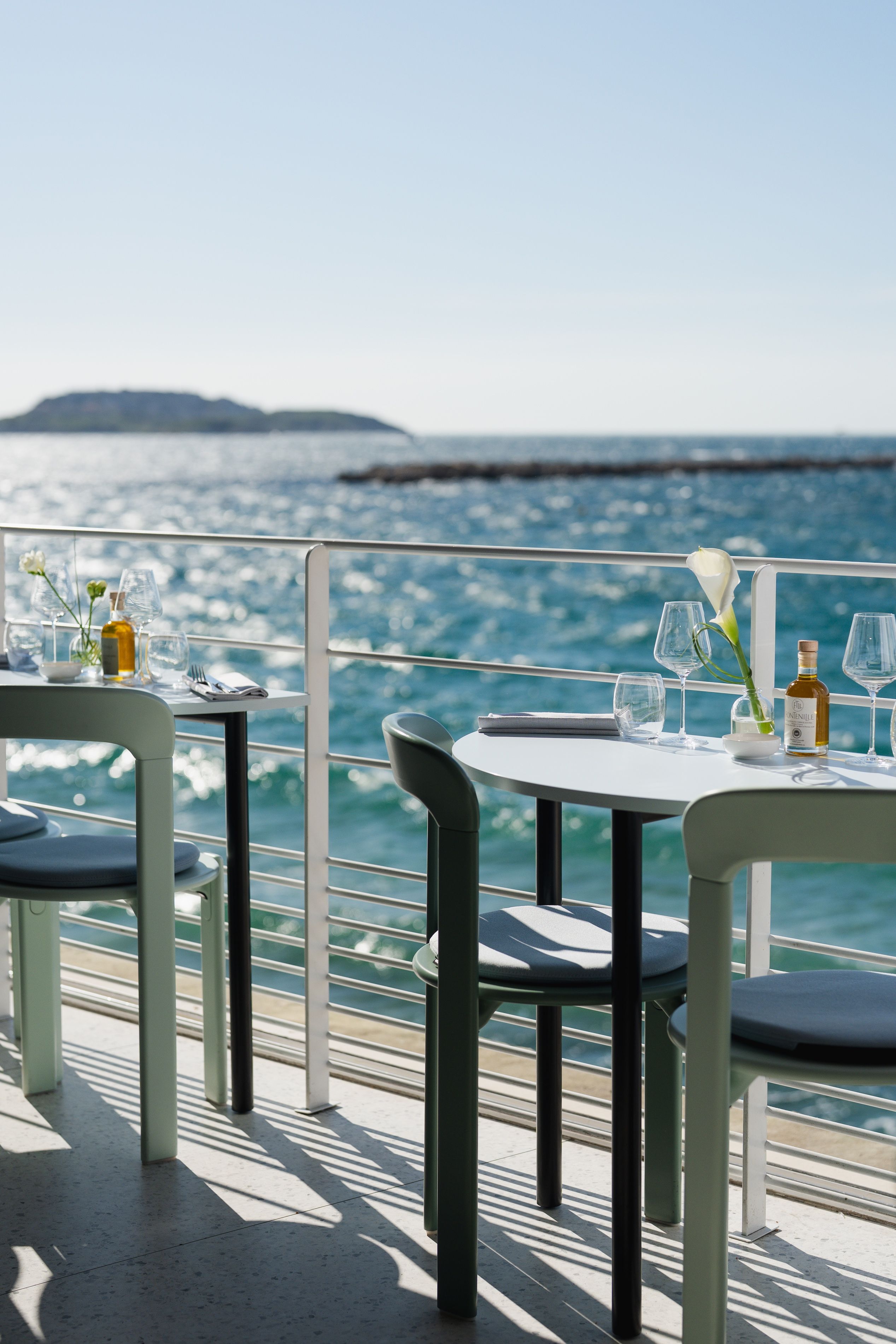 Un comedor con mesas y sillas frente a una masa de agua, con vista al mar y la costa distante.
