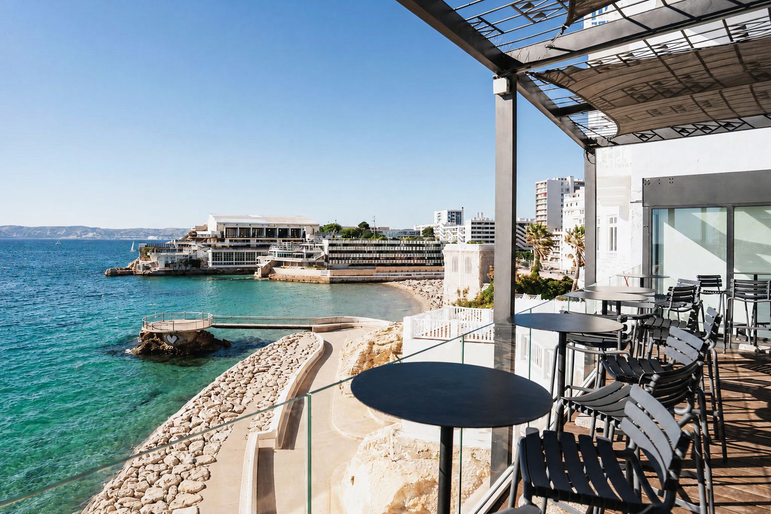 Una terraza frente al mar con asientos al aire libre, vista al océano y edificios en el fondo.