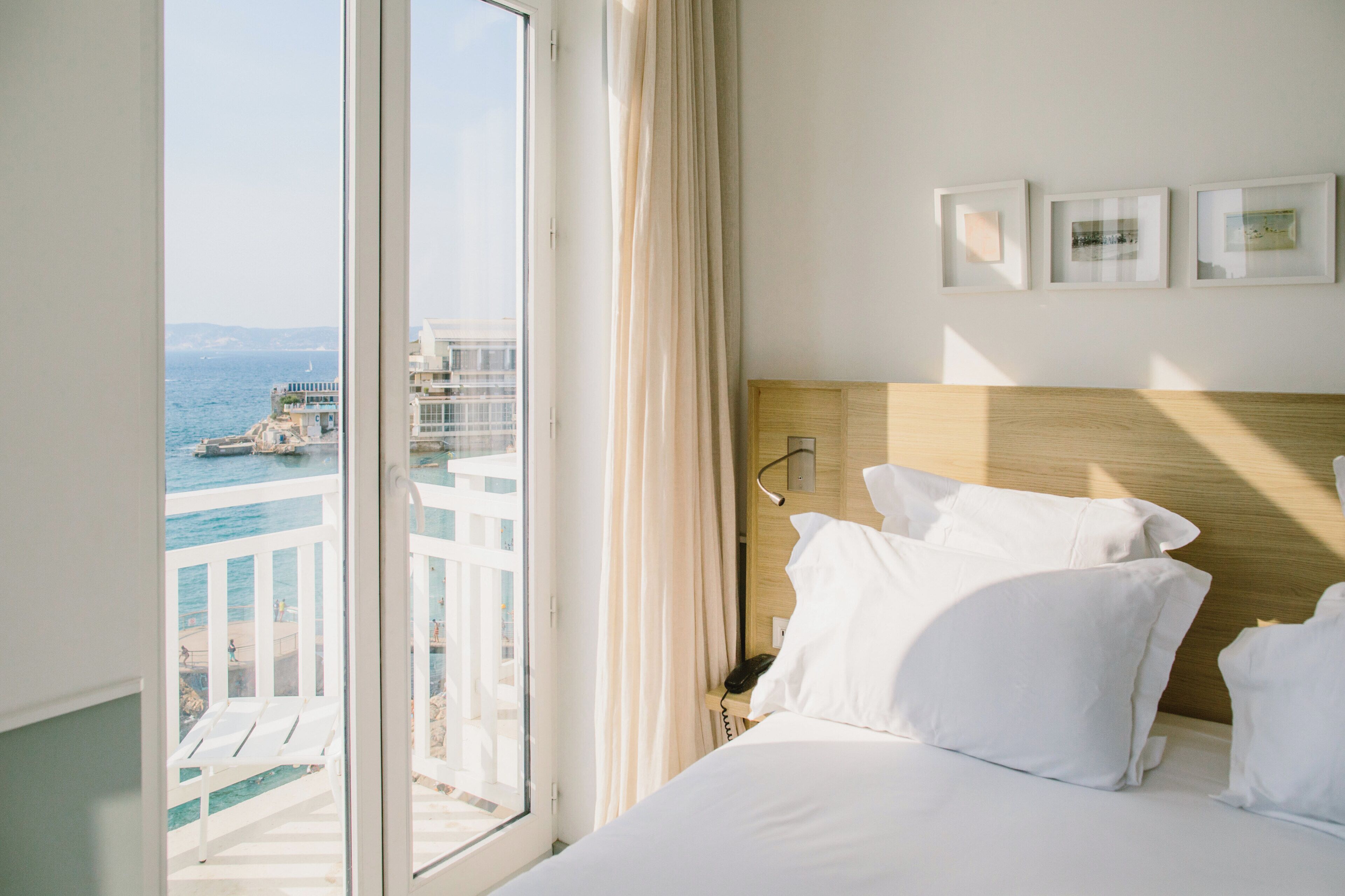 Quarto de hotel com uma janela grande, uma cama com lençóis brancos, cabeceira de madeira e três quadros na parede.