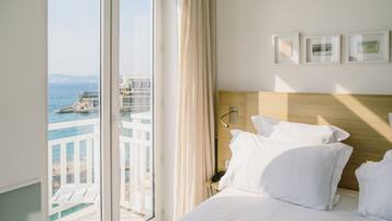 Quarto de hotel com uma janela grande, uma cama com lençóis brancos, cabeceira de madeira e três quadros na parede.