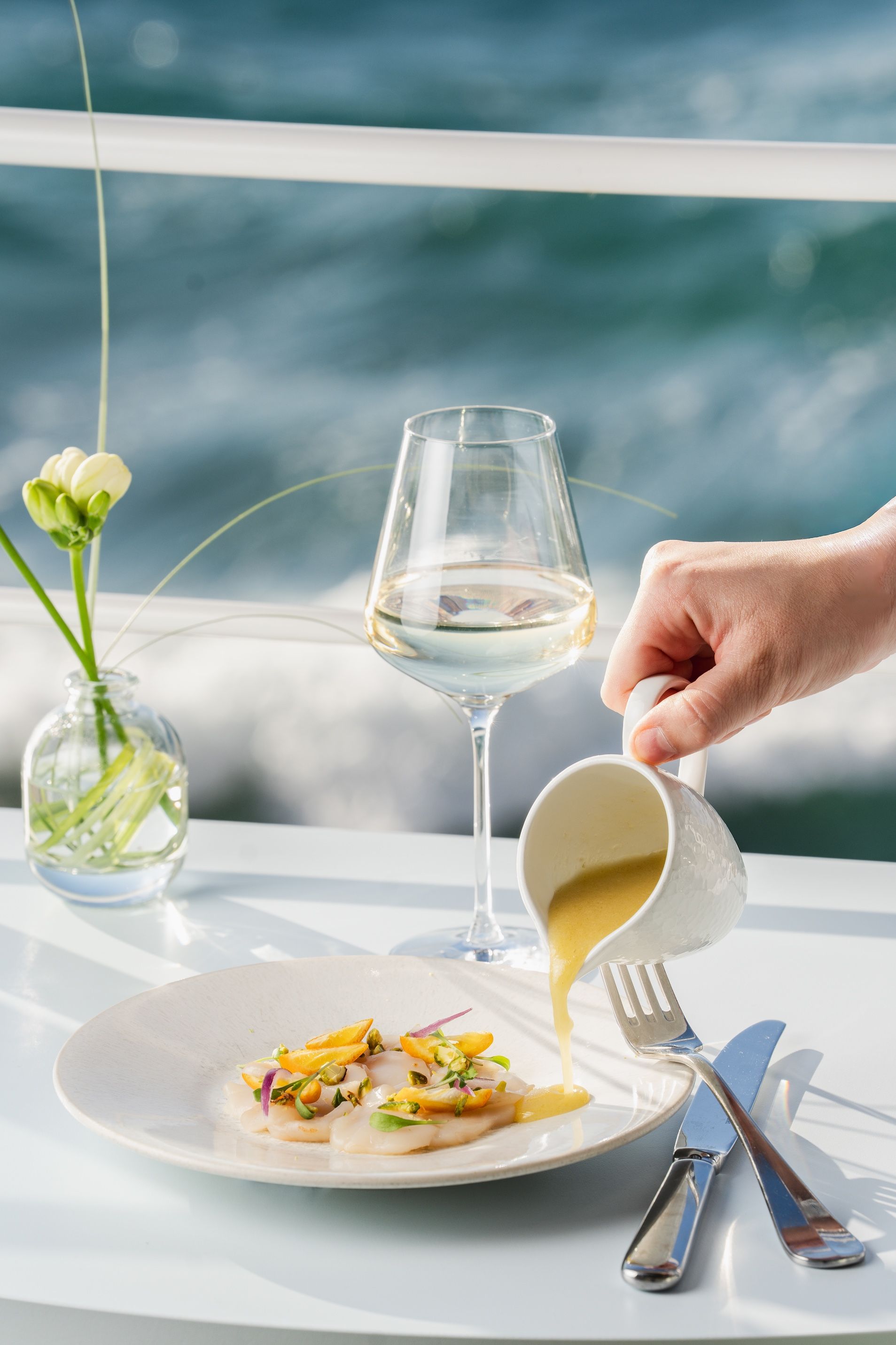 A dining table with a plate of food, a glass of white wine, and a hand pouring a yellow sauce.