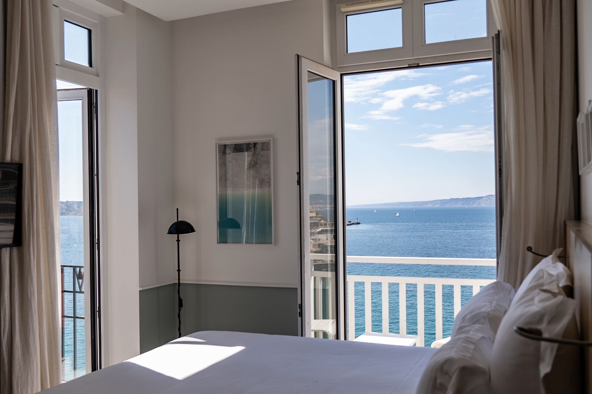 Quarto com cama, luminária de chão, pintura e vista para o mar.