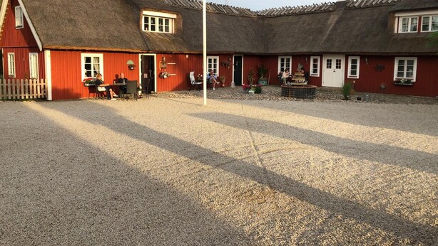 A red barn with a thatched roof, a gravel driveway, and a small outdoor seating area with tables and chairs. - Sanddala Bed & Breakfast (Everöd)