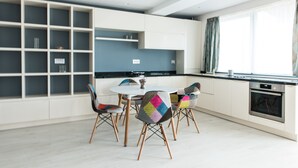 Una cocina moderna con armarios blancos, encimera negra y horno empotrado. Hay una mesa de comedor con sillas de colores y una ventana con cortinas.