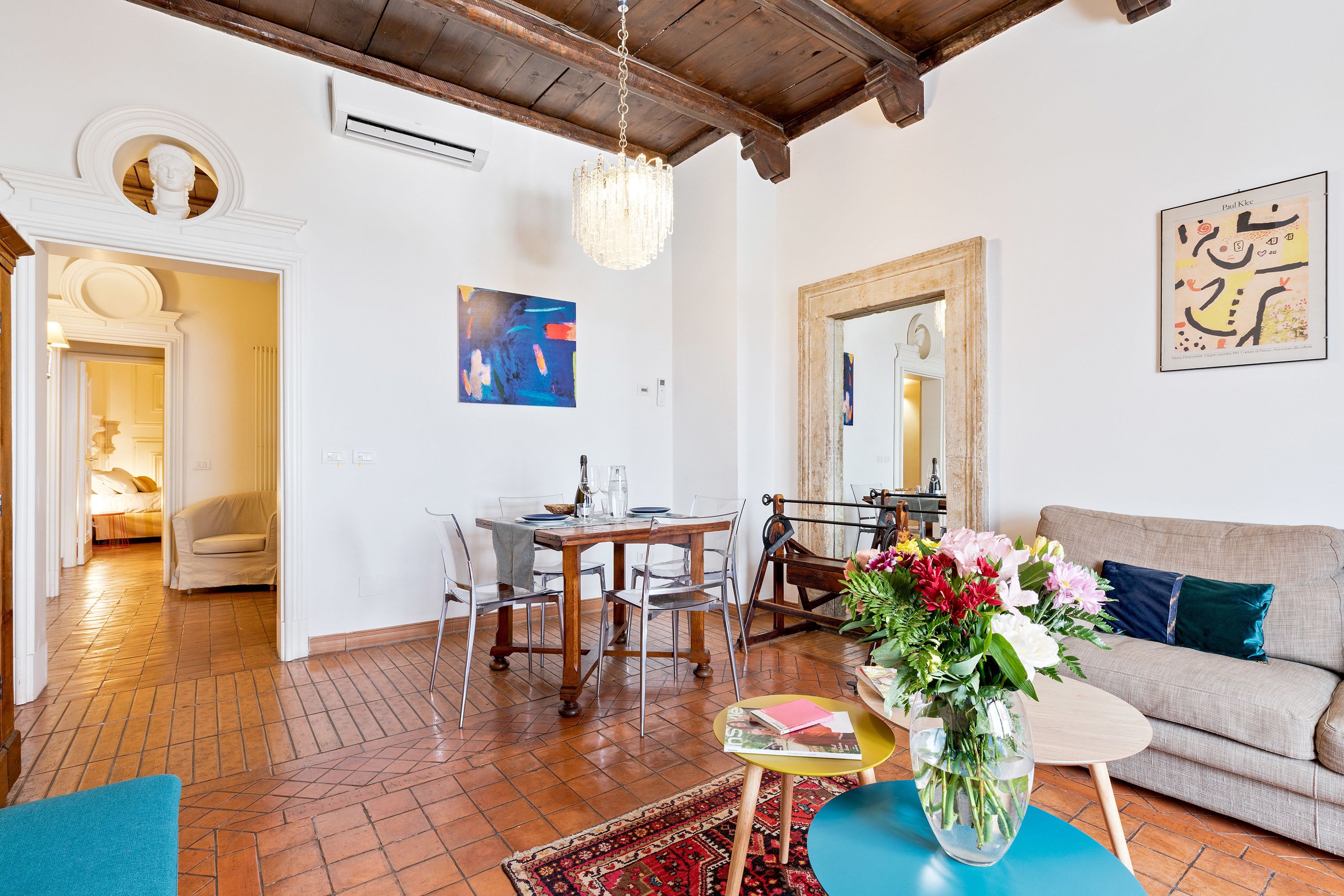 A living room with a sofa, a dining table with chairs, a chandelier, and a vase of flowers.