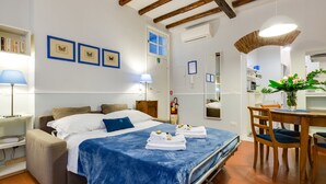 Une chambre bien éclairée, avec un lit, une table de chevet et un bureau avec des chaises. On y trouve des tableaux encadrés au mur, une fenêtre et un appareil de climatisation.