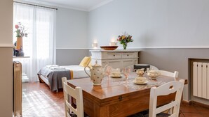 Une pièce avec une table à manger en bois, mise avec des tasses à thé et un vase de fleurs. On y trouve un lit avec une tête de lit blanche et une fenêtre ornée de rideaux légers.