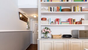 Une cuisine avec des meubles blancs, un four à micro-ondes et une bibliothèque remplie de livres et d’objets décoratifs.