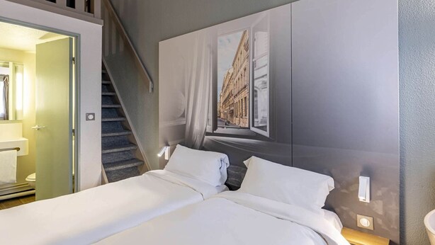 A hotel room with a bed, a painting of a building, a staircase, and a bathroom visible through an open door. - B&B Hotel Bordeaux Merignac Hotel De Ville (Merignac)