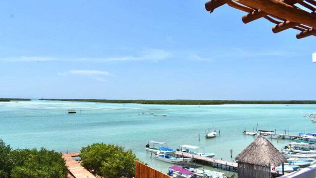 A coastal view with a wooden deck overlooking turquoise waters, boats docked at a pier, and a thatched-roof structure. - Hotel Rio Lagartos (Río Lagartos)
