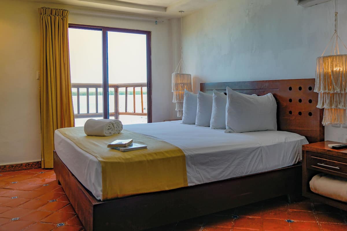 A hotel room with a large bed, a wooden headboard, a nightstand, a bedside table, a hanging light fixture, and a view of the outdoors through a window.