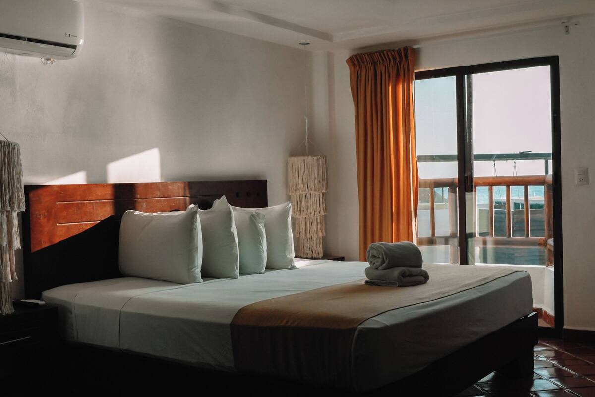 A hotel room with a bed, a nightstand, a hanging light fixture, a window with curtains, and a view of the outside.