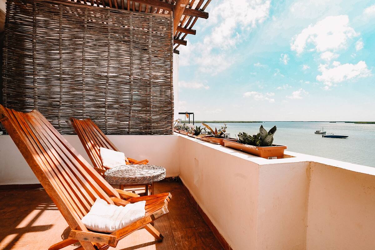 A balcony with two wooden lounge chairs, a wicker wall, and potted plants.