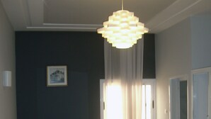 A bedroom with a bed, a bedside table, a hanging light fixture, a window with curtains, and a door leading to another room.