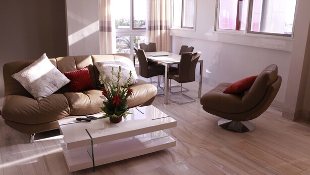 Ein modernes Wohnzimmer mit einem beigen Sofa, einem weißen Couchtisch und einem braunen Sessel. Im Hintergrund ist eine Essecke mit Tisch und Stühlen zu sehen.
