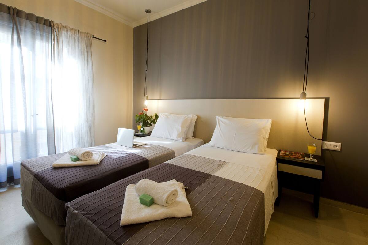 A hotel room with two beds, a bedside table with a lamp, a laptop, and a small potted plant.