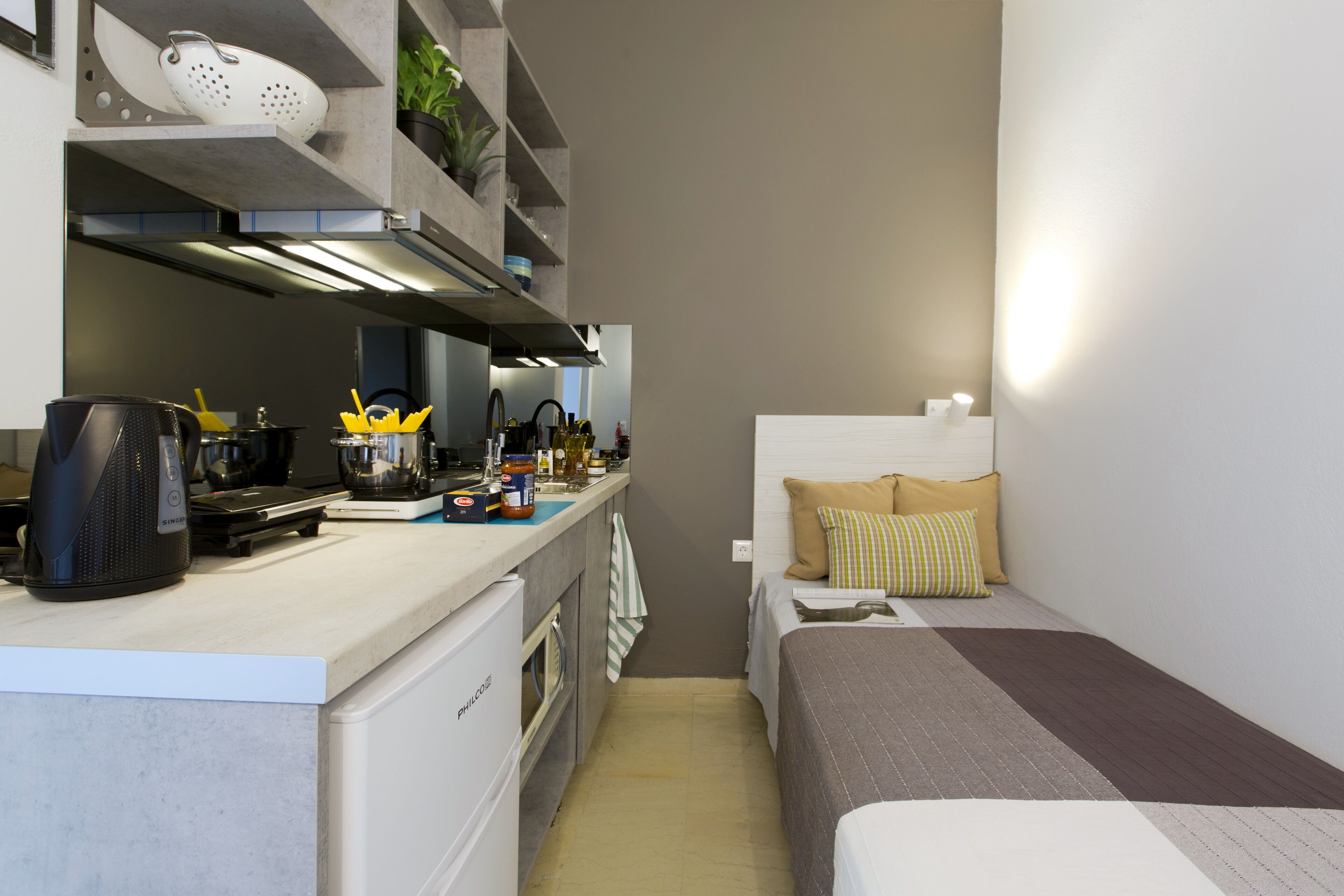 A compact kitchen with a coffee maker, kettle, and various kitchen items on the counter. A small refrigerator is visible on the left, and a bed with a pillow is on the right.