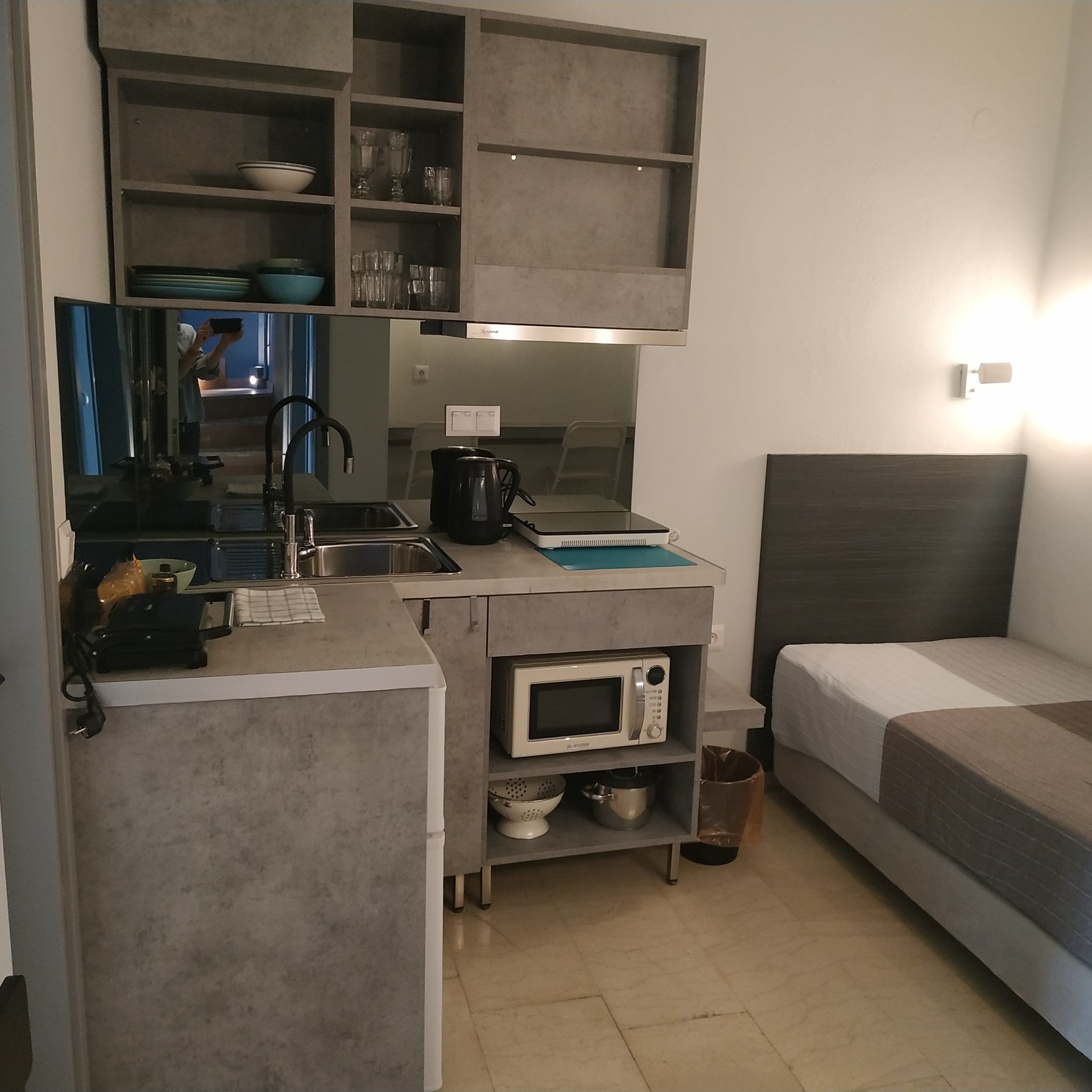 Een kleine keuken met een magnetron, waterkoker en wastafel. Een bed is rechts zichtbaar.