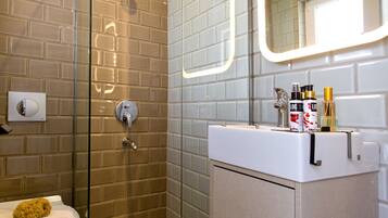 Un baño moderno con cabina de ducha de vidrio, un lavamanos blanco con artículos de aseo y un espejo con iluminación LED.