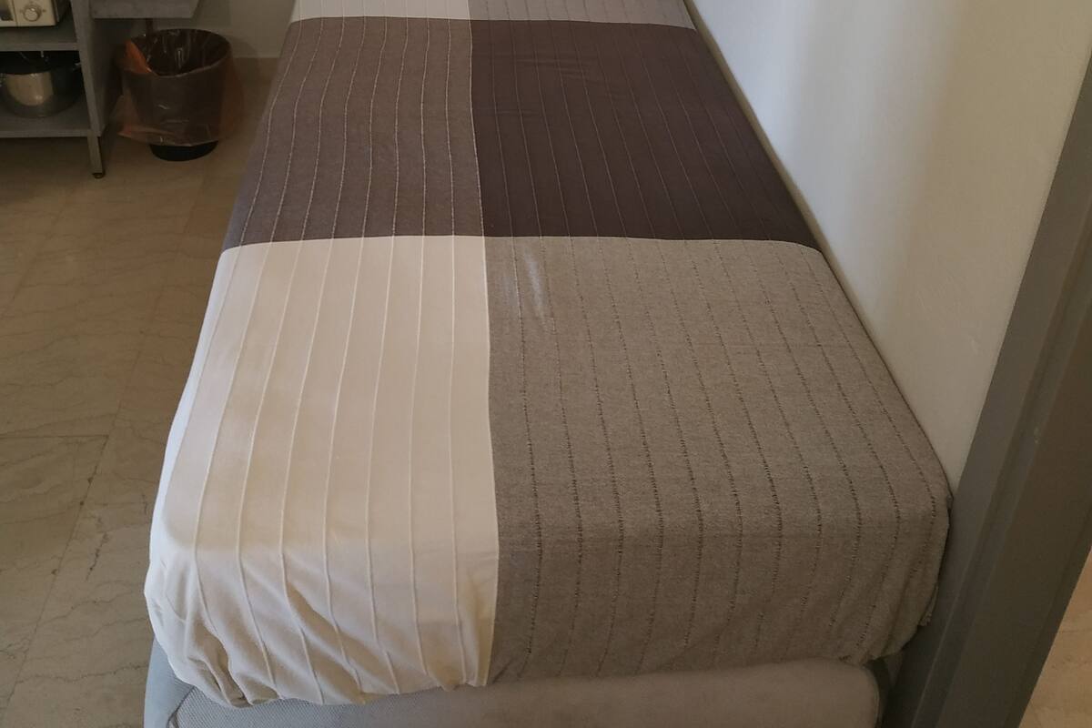 A small, single-bed room with a dark headboard, a bedside table with a lamp, and a wall-mounted light.