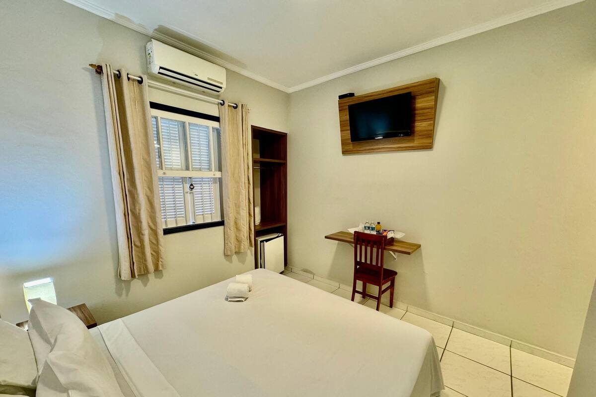 A hotel room with a bed, a desk with a chair, a TV mounted on the wall, and an air conditioning unit.