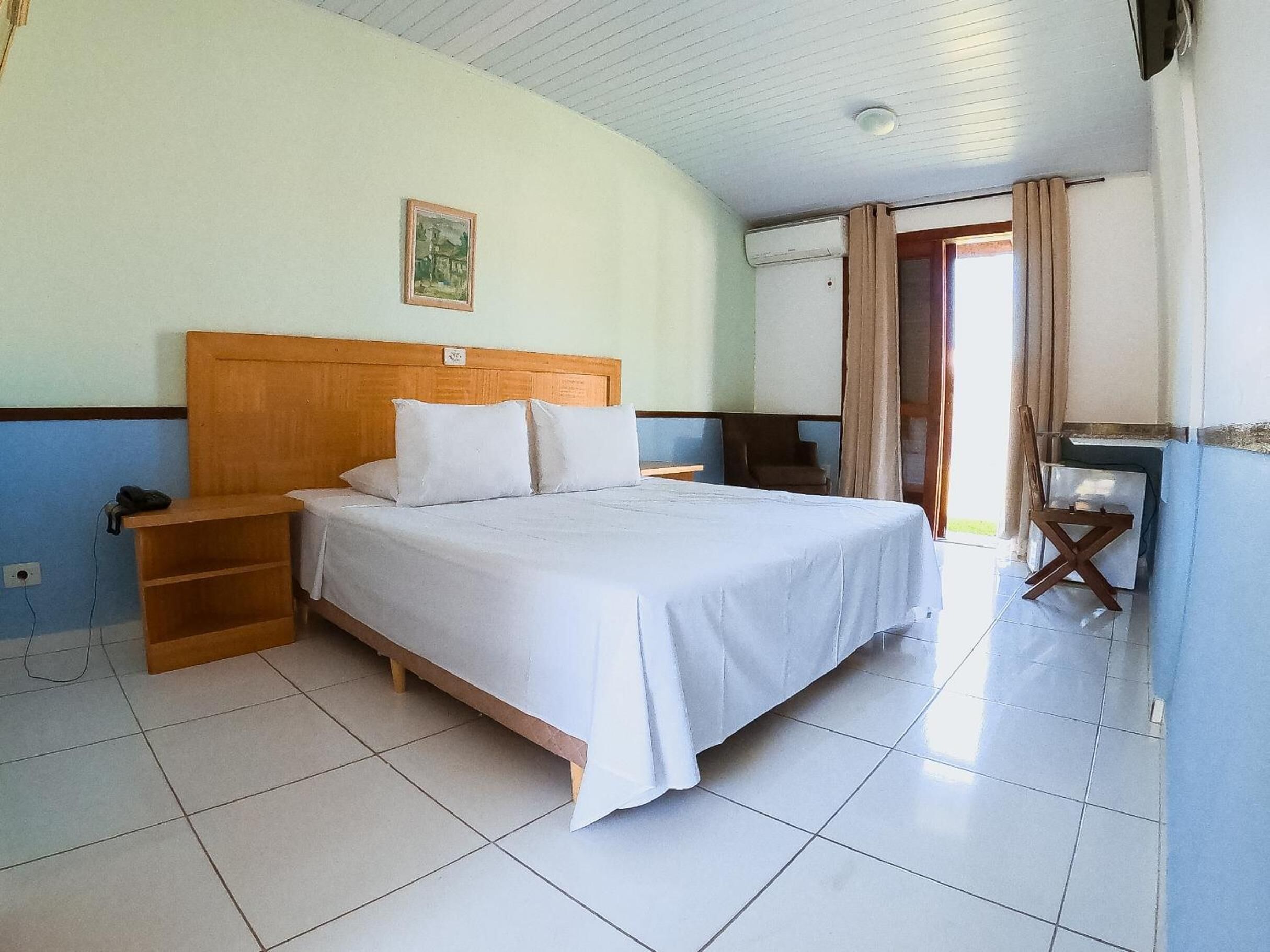 A hotel room with a wooden bed, white bedding, a small bedside table, a chair, and a TV.