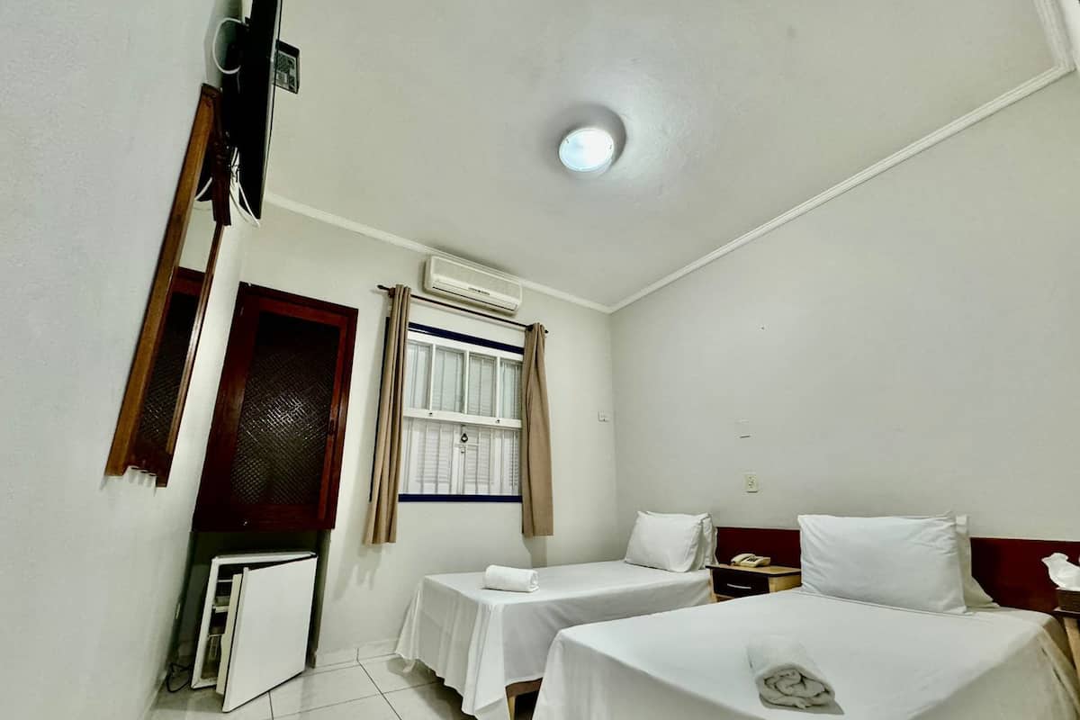 A hotel room with two single beds, a small table, a desk with a telephone, and an air conditioning unit.