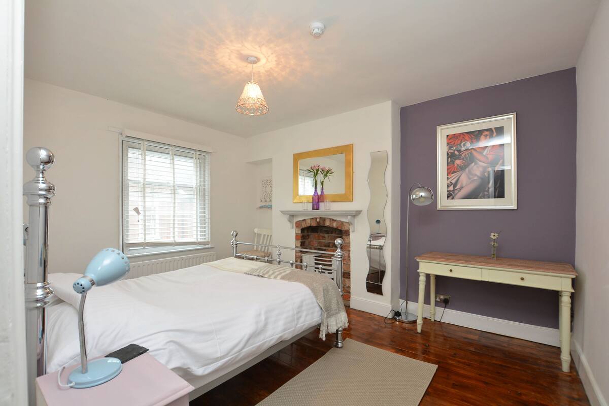 Quarto com cama, luminária de cabeceira, cômoda e lareira.