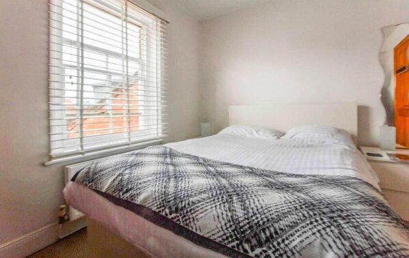 A bedroom with a bed, a window with blinds, a nightstand, and a lamp.