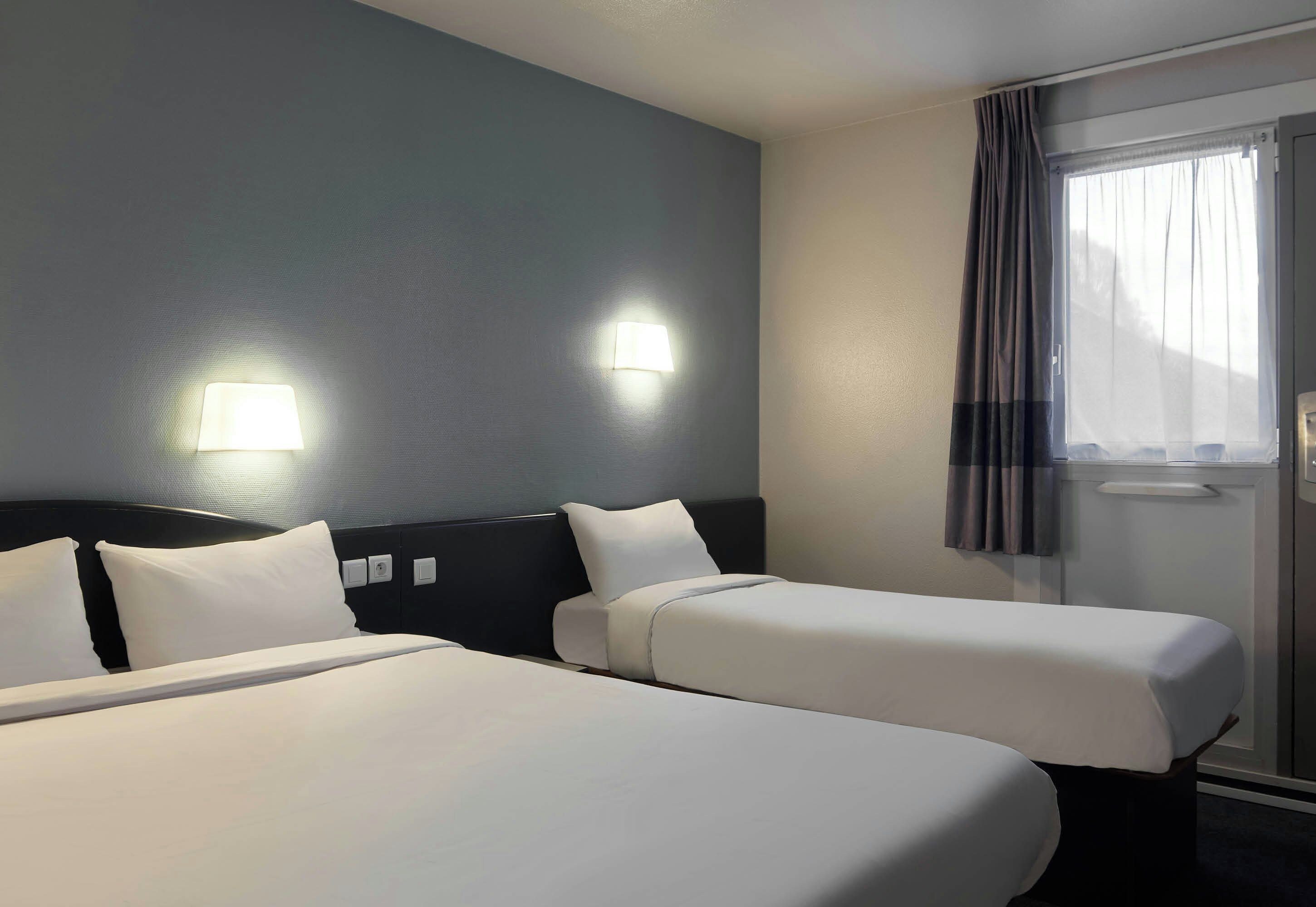 A hotel room with two beds, a headboard, a window with curtains, and wall-mounted lights.