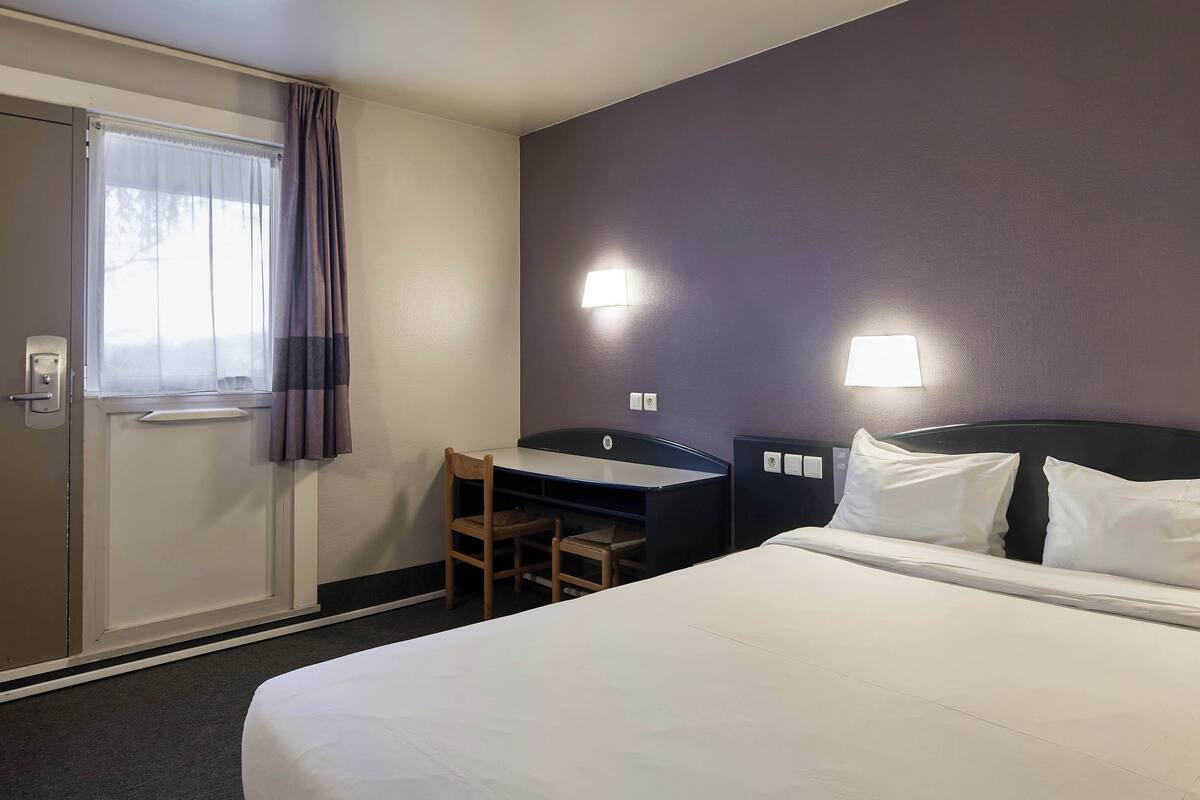 Habitación de hotel con cama, escritorio, silla, ventana con cortinas y dos lámparas de pared.