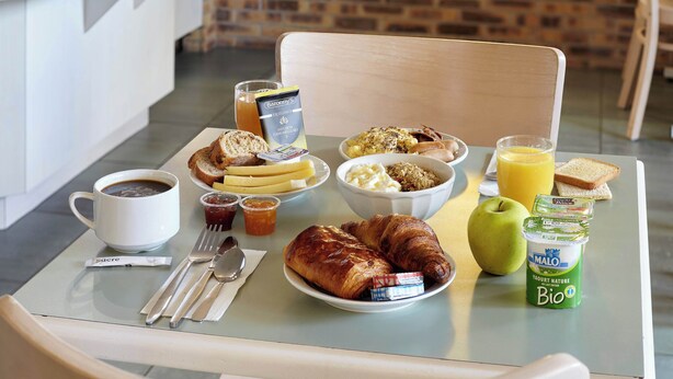 Et morgenbord med croissanter, brød, smør, marmelade, en skål müsli, et glas appelsinjuice, en kop kaffe og en mælkeflaske.