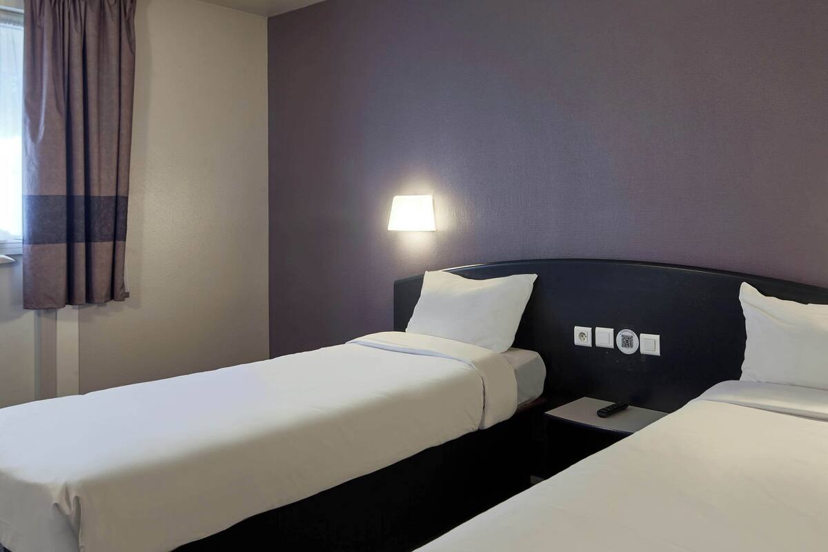 Habitación de hotel con dos camas individuales, una mesita de noche, una lámpara y una ventana con cortinas.