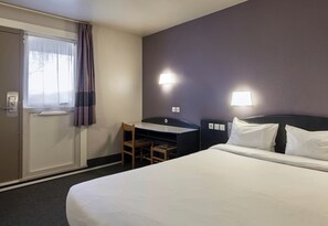 Une chambre d’hôtel comprenant un lit, un bureau, une chaise, une fenêtre avec des rideaux et deux lampes fixées au mur.