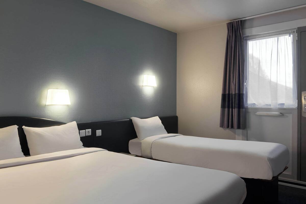 Habitación de hotel con dos camas, cabecera oscura, ventana con cortinas e iluminación en la pared.