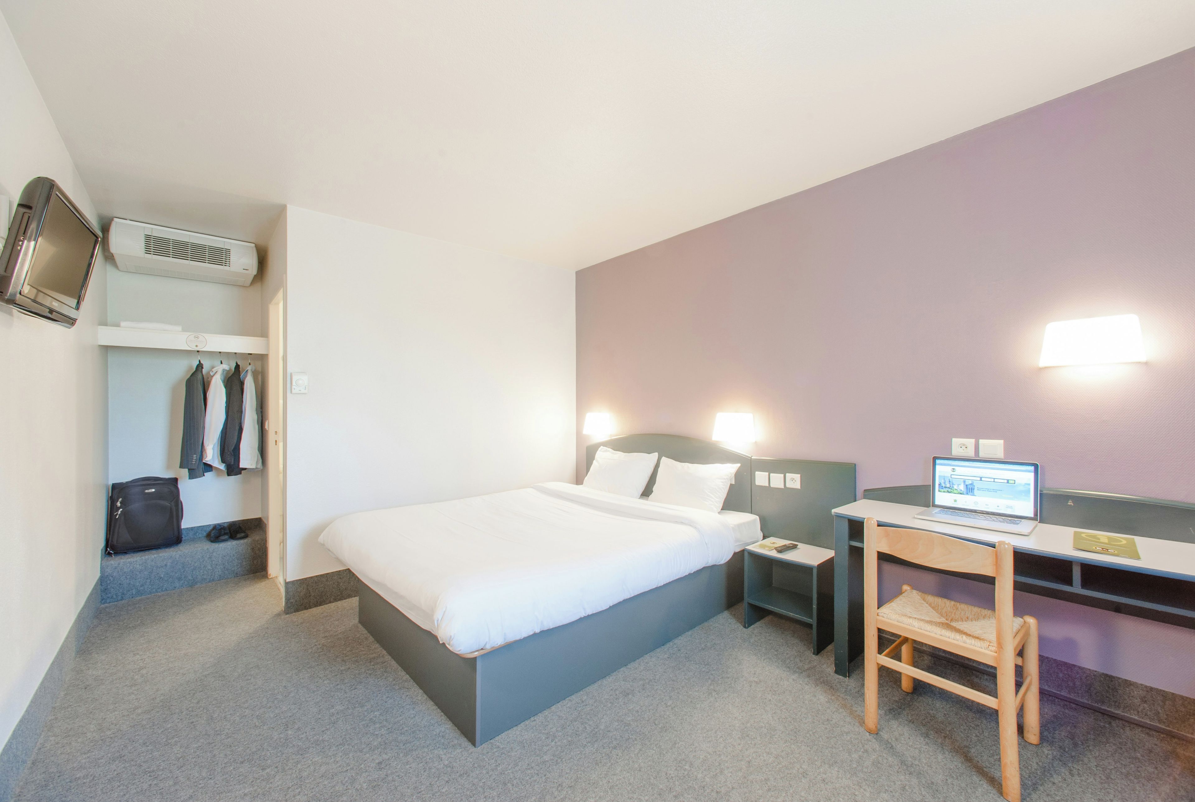 Habitació d'hotel amb un llit, una escriptori amb un ordinador portàtil, una cadira, un penjallabecs amb roba i un aparell d'aire condicionat.