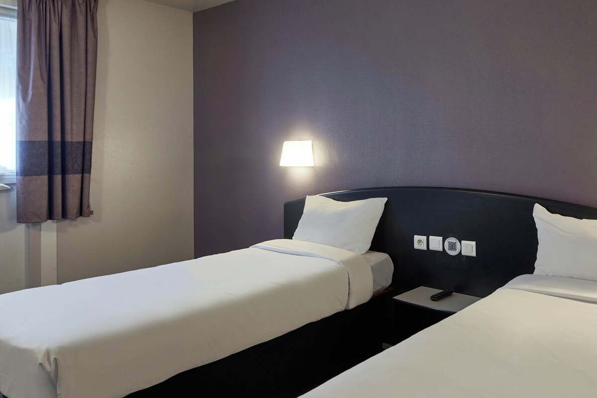 Habitación de hotel con dos camas individuales, una mesita de noche, una lámpara y una ventana con cortinas.