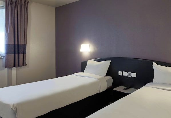 A hotel room with two single beds, a nightstand, a lamp, and a window with curtains.