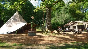Un entorno rústico al aire libre con un gran árbol, una estructura de madera que dice 'Woodland Cafe' y una tienda de campaña con luces colgantes.