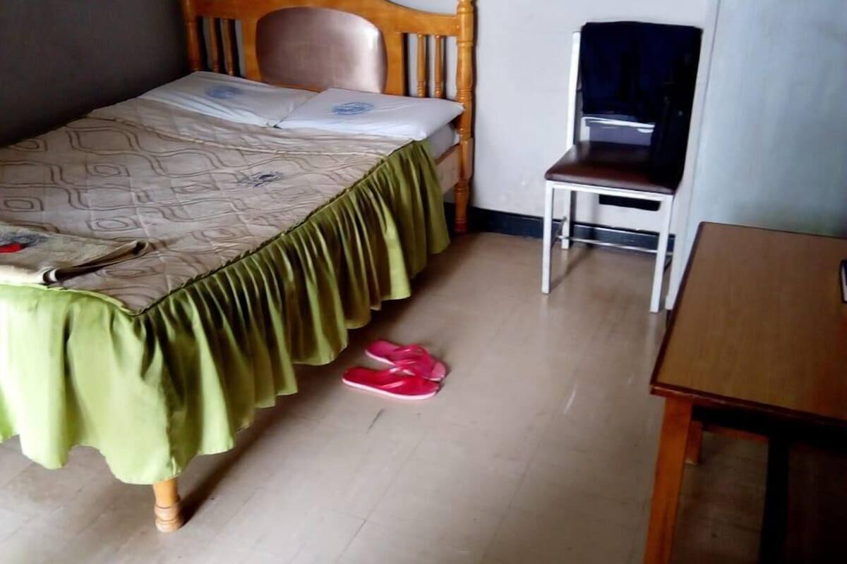 A bedroom with a wooden bed, a green bedspread, a pair of red slippers, a wooden nightstand, and a television on a stand.