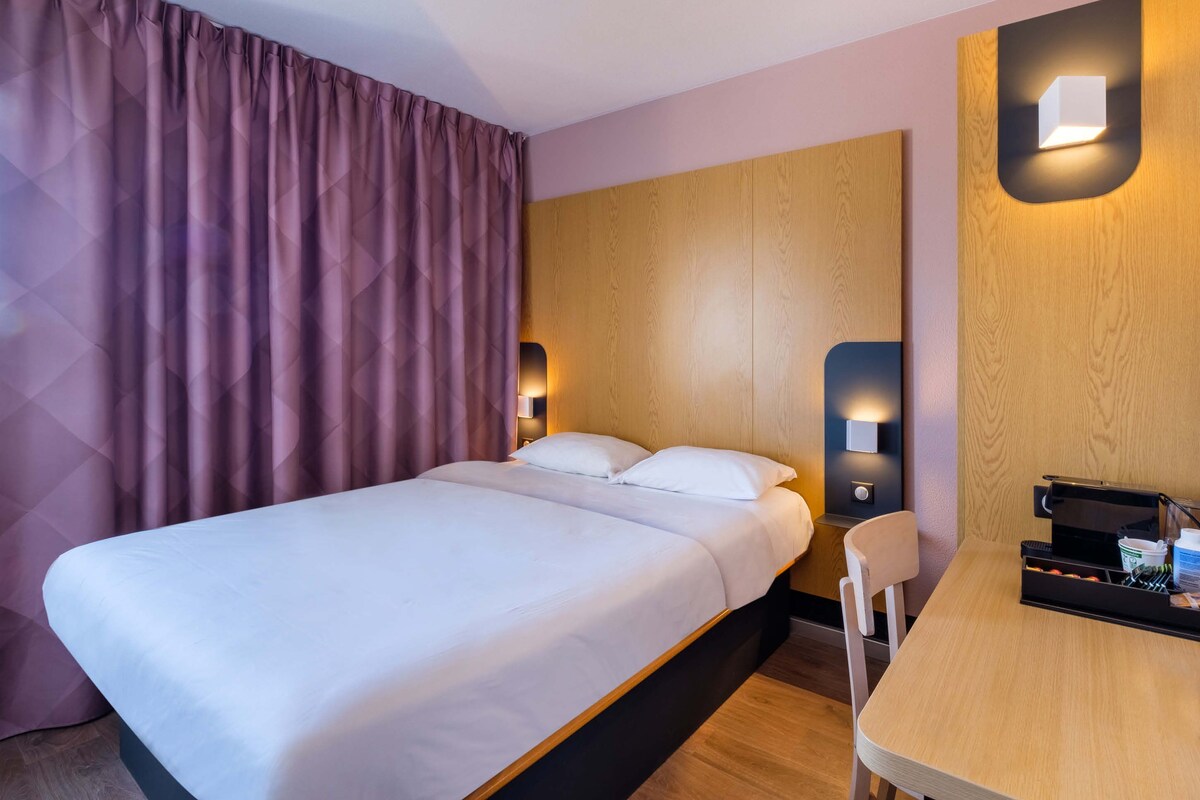 A hotel room with a bed, a wooden nightstand, a chair, and a desk with a coffee machine.