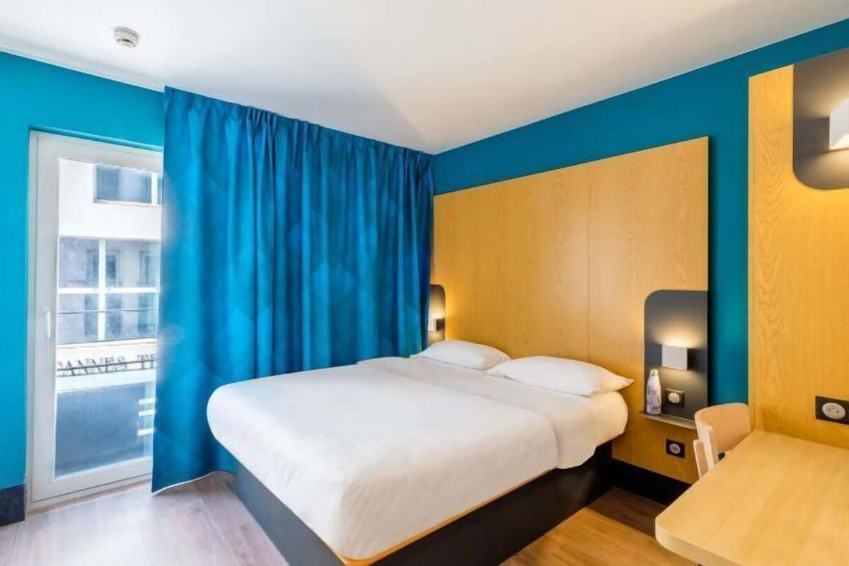 Une chambre d’hôtel avec un lit, un bureau, une chaise, une fenêtre avec des rideaux et une vue sur l’extérieur.