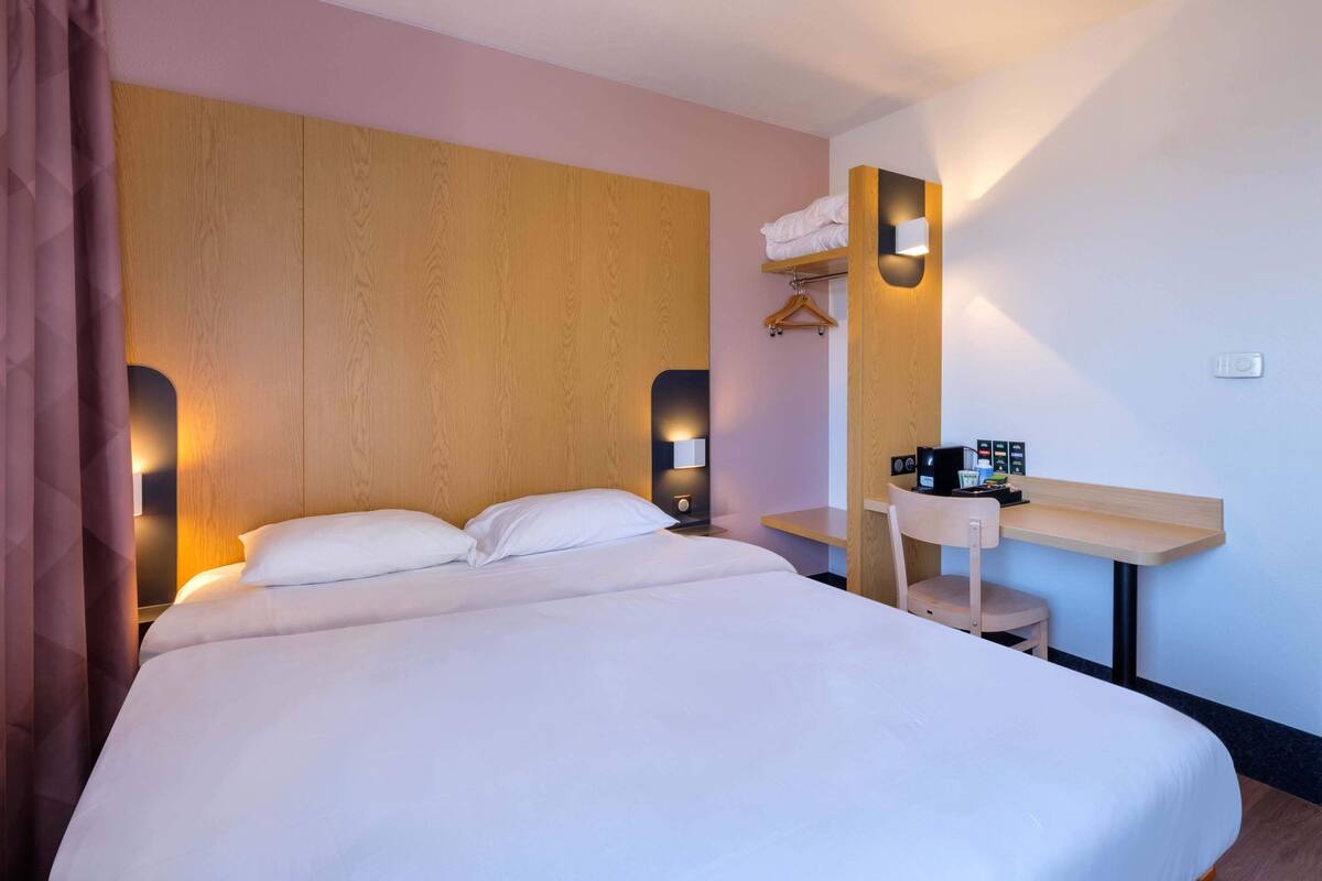 Une chambre d’hôtel avec un grand lit, une tête de lit en bois, des lampes de chevet, un petit bureau avec une chaise et une étagère murale où sont rangées les serviettes.