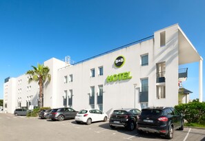 A modern hotel with a white facade, a prominent 'HOTEL' sign, and a parking lot with several cars. - B&B Hotel Cannes La Bocca Plage (Cannes)