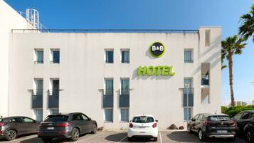 A modern hotel building with a sign reading "B&B HOTEL" and multiple parking spaces in front.