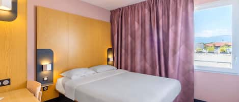 A hotel room with a bed, a desk, and a window with curtains.