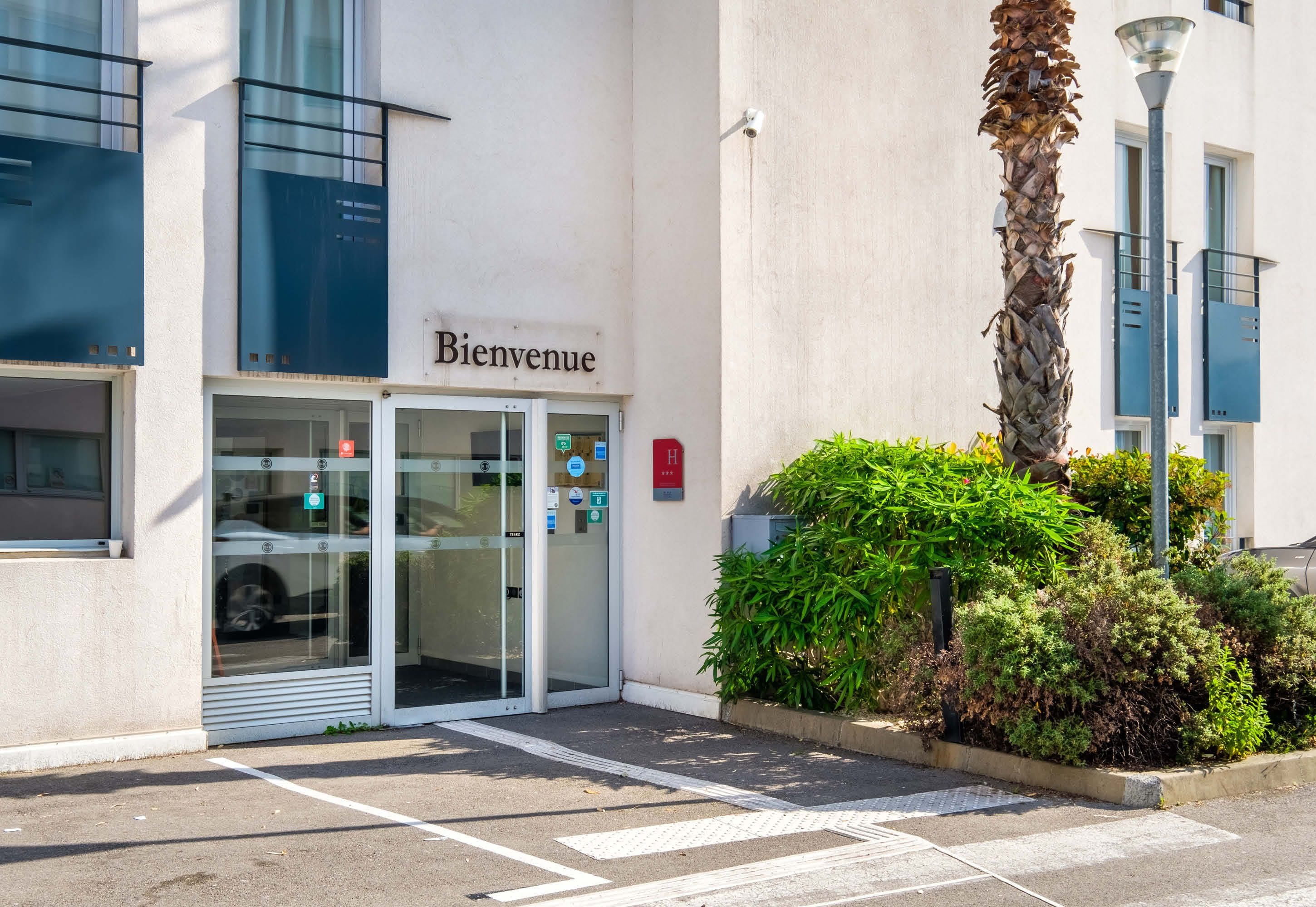A hotel entrance with a glass door, a 'Bienvenue' sign, and a parking area.