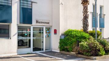 A hotel entrance with a glass door, a 'Bienvenue' sign, and a parking area.