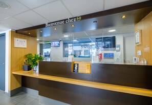 Un comptoir d’accueil avec une pancarte « Bienvenue chez… », une plante en pot et un écran numérique.